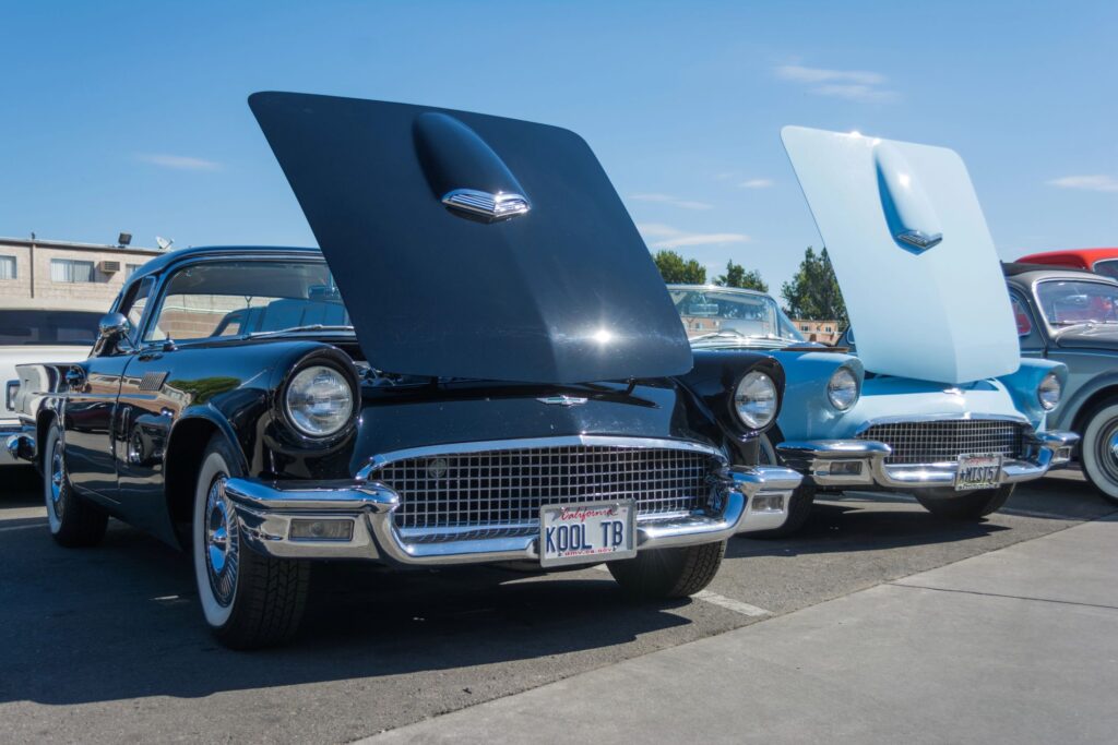A line of classic 1957 Ford Thunderbirds in black, light blue, and grey with their hoods open at a classic car auction