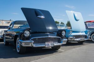 A line of classic 1957 Ford Thunderbirds in black, light blue, and grey with their hoods open at a classic car auction