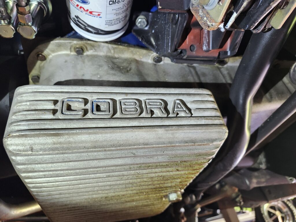 Under-vehicle view of a ribbed aluminum "Cobra" branded oil pan on a Ford small-block engine.