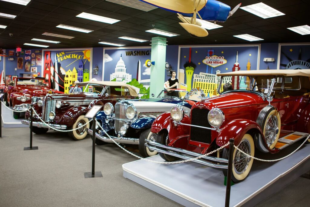 A line of pre-war and mid-century luxury sedans, including Buicks and Duesenbergs, in a museum with colorful wall murals.