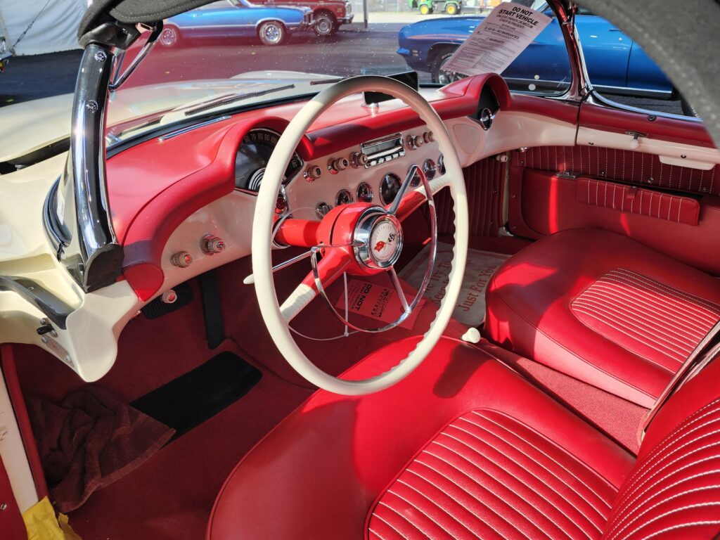 Interior view of a 1953 Corvette featuring red upholstery, a two-spoke steering wheel, and black dial gauges.