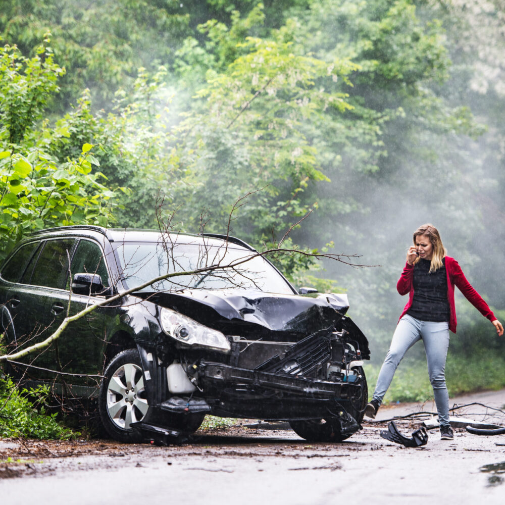 An angry and frustrated young woman with smartphone by the damaged car after a car accident, making a phone call. Copy space.